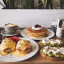 A photo of a brunch spread with eggs benedict, pancakes, and avocado toast. There's a coffee cup with a saucer and a few utensils. The brunch is placed on a wooden board. In the background, there's a white wall with a few potted plants.