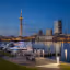 A photo of the Southampton city skyline at night with the landmark Spinnaker Tower shining brightly. There are boats docked along the waterfront. In the foreground, there's a wooden deck with chairs, and a lamp post. There's a path leading to the deck. The water reflects the city skyline. The background has a few buildings.