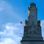The image shows a monument with birds perched on it against a blue sky. The composition is decent, with the monument centered. The lighting is good, with clear daylight. Sharpness is acceptable, allowing for some detail to be seen in the stone carvings. Color accuracy appears good, with the blue sky and the stone's natural color represented well. The image is reasonably photo-realistic. The key elements of the monument are in focus. The image is well-composed, well-lit, and sharp enough to convey the details of the monument. There are no unwanted elements. The birds add an interesting element to the photo.