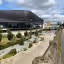 The image showcases a modern building exterior with a plaza in front. The composition is decent, capturing the building and its surroundings. The lighting is good, with natural light and some cloud cover. The sharpness is acceptable, though not exceptionally crisp. The colors appear accurate. The image is reasonably photo-realistic. Key elements, such as the building's facade and the plaza's layout, are in focus. The image is well-composed with good use of leading lines. The image is well-lit and the colors are accurate. The image is sharp enough to see the details of the building and the plaza. The image is photo-realistic and the key elements are in focus.