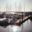 The image shows a marina filled with boats and sailboats. The composition is decent, but the lighting is a bit harsh due to the sun reflecting off the water, which slightly impacts the overall quality. Sharpness is acceptable, but not exceptional. Color accuracy seems fine. The image is reasonably photo-realistic. Key elements are generally in focus. It's a bit cluttered, but that's inherent to the scene. The sun glare is a distraction. Considering the criteria, the image scores as follows: Well-composed (20/25), Well-lit (10/15), Sharp (15/20), Color-accurate (8/10), Photo-realistic (12/20), Key elements in focus (10/10).